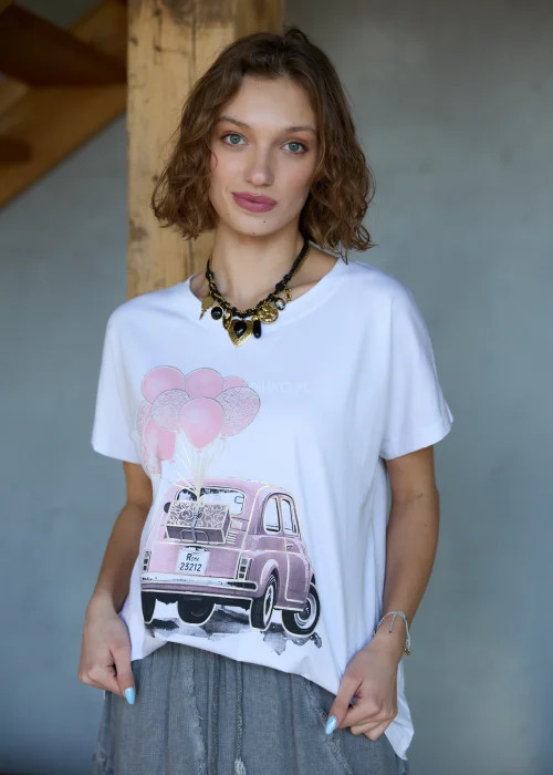 Biała bluzka damska z nadrukiem różowego auta i balonów, o luźnym kroju, stylizowana z szarymi spodniami i biżuterią.