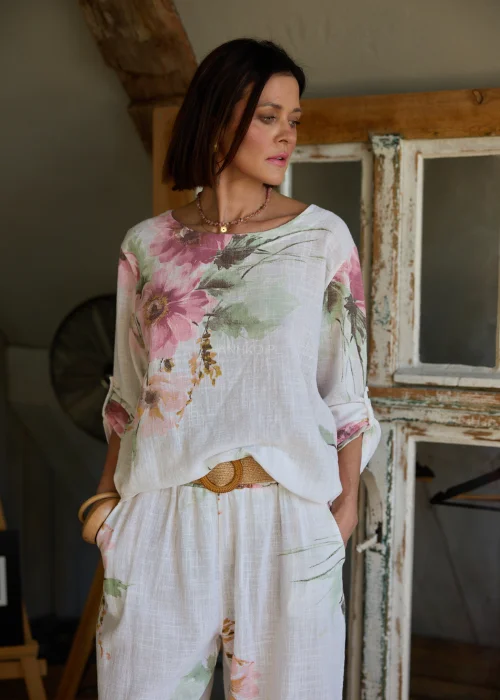Bluzka damska w kolorze białym z delikatnym kwiatowym printem, luźny fason z podwijanymi rękawami, stylizowana z szerokimi spodniami i paskiem w naturalnym, eleganckim looku.