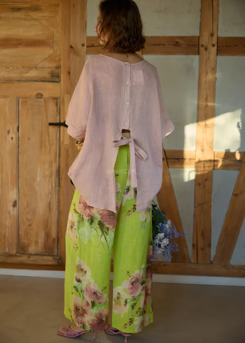 Różowa bluzka lniana damska o luźnym kroju z krótkim rękawem i wiązaniem z tyłu, zestawiona z zielonymi spodniami w kwiaty w naturalnej stylizacji.