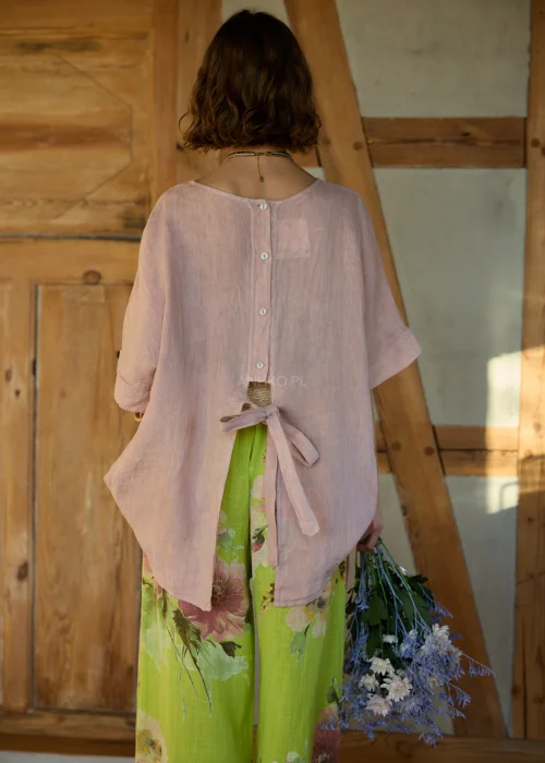 Różowa bluzka lniana damska o luźnym kroju z krótkim rękawem i wiązaniem z tyłu, zestawiona z zielonymi spodniami w kwiaty w naturalnej stylizacji.