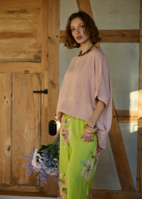 Różowa bluzka lniana damska o luźnym kroju z krótkim rękawem i wiązaniem z tyłu, zestawiona z zielonymi spodniami w kwiaty w naturalnej stylizacji.
