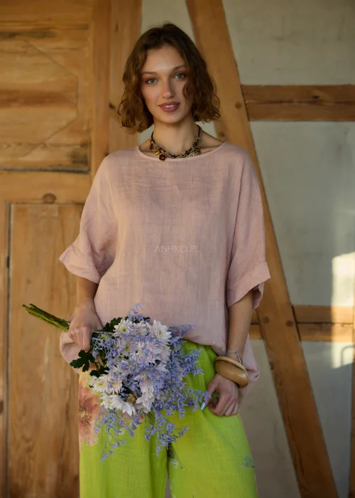 Różowa bluzka lniana damska o luźnym kroju z krótkim rękawem i wiązaniem z tyłu, zestawiona z zielonymi spodniami w kwiaty w naturalnej stylizacji.
