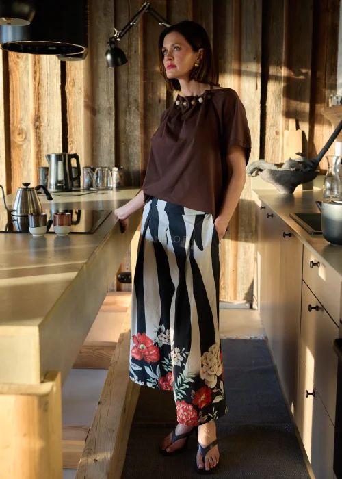 Brązowa bluzka damska o luźnym kroju z ozdobnym dekoltem i krótkim rękawem, zestawiona ze spodniami w kontrastowy wzór w naturalnym wnętrzu.