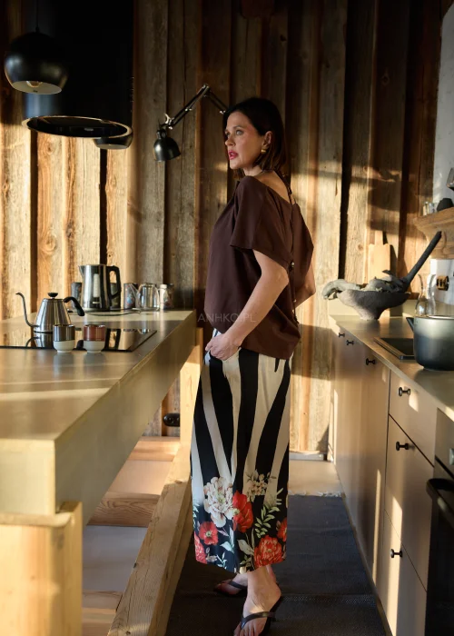 Brązowa bluzka damska o luźnym kroju z ozdobnym dekoltem i krótkim rękawem, zestawiona ze spodniami w kontrastowy wzór w naturalnym wnętrzu.