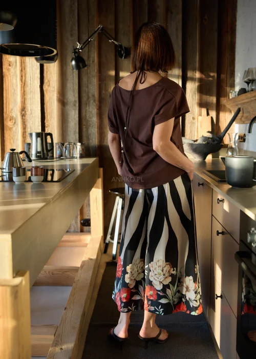 Brązowa bluzka damska o luźnym kroju z ozdobnym dekoltem i krótkim rękawem, zestawiona ze spodniami w kontrastowy wzór w naturalnym wnętrzu.