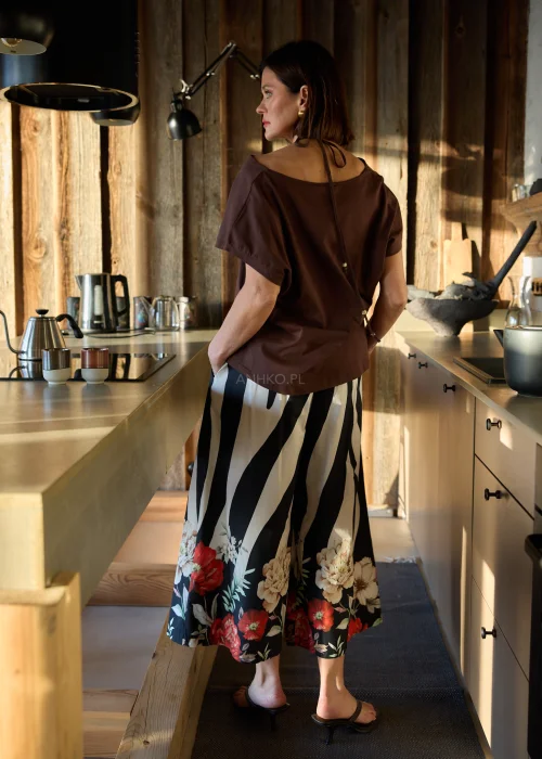 Brązowa bluzka damska o luźnym kroju z ozdobnym dekoltem i krótkim rękawem, zestawiona ze spodniami w kontrastowy wzór w naturalnym wnętrzu.
