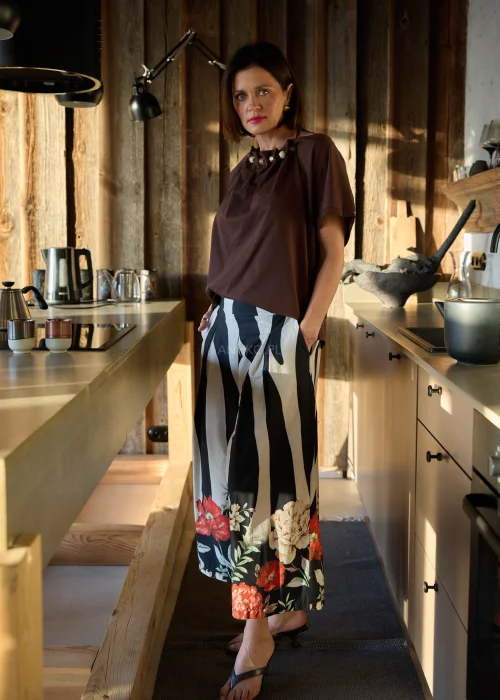 Brązowa bluzka damska o luźnym kroju z ozdobnym dekoltem i krótkim rękawem, zestawiona ze spodniami w kontrastowy wzór w naturalnym wnętrzu.