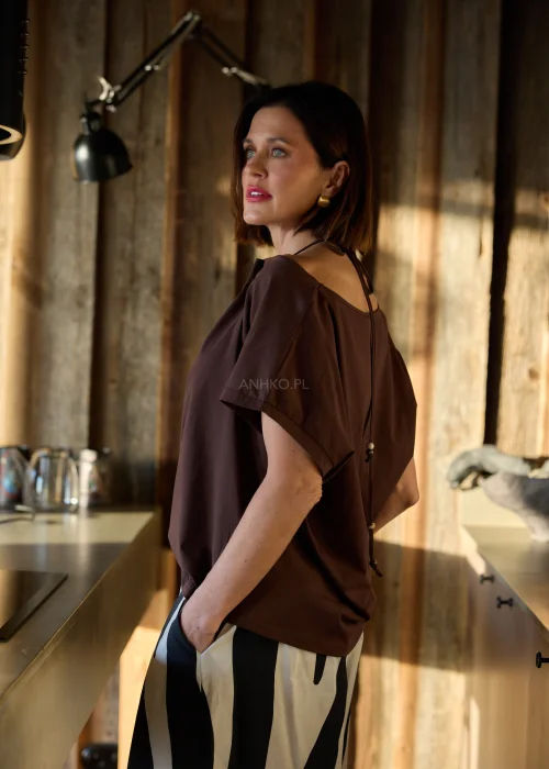 Brązowa bluzka damska o luźnym kroju z ozdobnym dekoltem i krótkim rękawem, zestawiona ze spodniami w kontrastowy wzór w naturalnym wnętrzu.