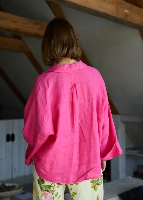 Lniana koszula oversize w odcieniu magenta z długim rękawem i guzikami, o luźnym fasonie, zestawiona z jasnymi spodniami, stylizacja w naturalnym, minimalistycznym klimacie.