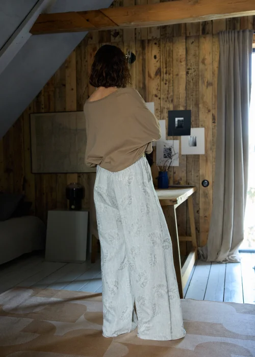 Lniane spodnie w szarym kolorze z subtelnym roślinnym wzorem, o szerokiej nogawce i luźnym kroju, stylizowane z paskiem w naturalnym odcieniu w minimalistycznym, eleganckim looku.