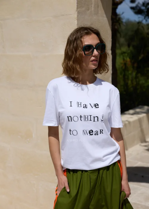 Biały T-shirt damski z nadrukiem „I Have Nothing To Wear” w czarnym kolorze. Luźny model casual zestawiony z zielonymi spodniami i okularami przeciwsłonecznymi.