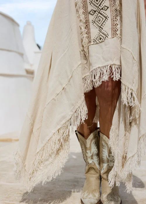 Sukienka damska maxi  w kolorze ecru bez ramiączek z etnicznym wzorem i dekoracyjnymi frędzlami. Zwiewny fason w stylu boho pięknie układa się na sylwetce, tworząc efektowną i kobiecą stylizację.