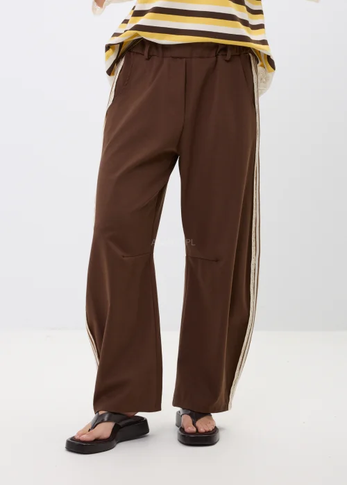 Brązowe spodnie z szeroką nogawką i ozdobnymi lampasami po bokach. Komfortowy fason z kieszeniami oraz kontrastowymi paskami i koronkowym wykończeniem nadaje stylizacji sportowej elegancji.