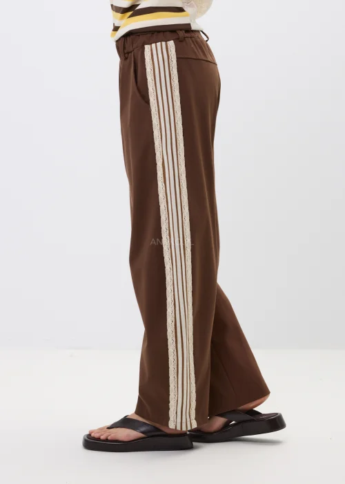 Brązowe spodnie z szeroką nogawką i ozdobnymi lampasami po bokach. Komfortowy fason z kieszeniami oraz kontrastowymi paskami i koronkowym wykończeniem nadaje stylizacji sportowej elegancji.