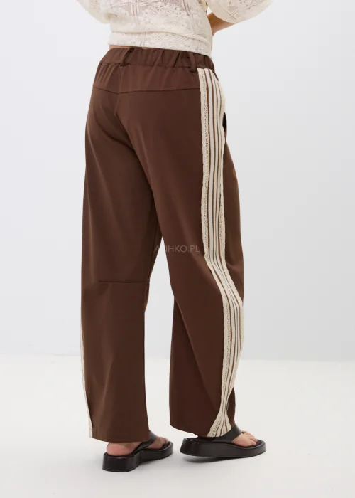 Brązowe spodnie z szeroką nogawką i ozdobnymi lampasami po bokach. Komfortowy fason z kieszeniami oraz kontrastowymi paskami i koronkowym wykończeniem nadaje stylizacji sportowej elegancji.