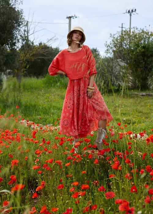 Koralowa sukienka maxi na cienkich ramiączkach z ozdobnym wzorem paisley. Zwiewny model o rozkloszowanym kroju prezentowany w stylizacji boho z kapeluszem i kowbojkami.