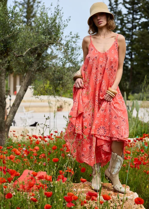 Koralowa sukienka maxi na cienkich ramiączkach z ozdobnym wzorem paisley. Zwiewny model o rozkloszowanym kroju prezentowany w stylizacji boho z kapeluszem i kowbojkami.