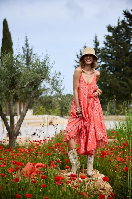 Koralowa sukienka maxi na cienkich ramiączkach z ozdobnym wzorem paisley. Zwiewny model o rozkloszowanym kroju prezentowany w stylizacji boho z kapeluszem i kowbojkami.
