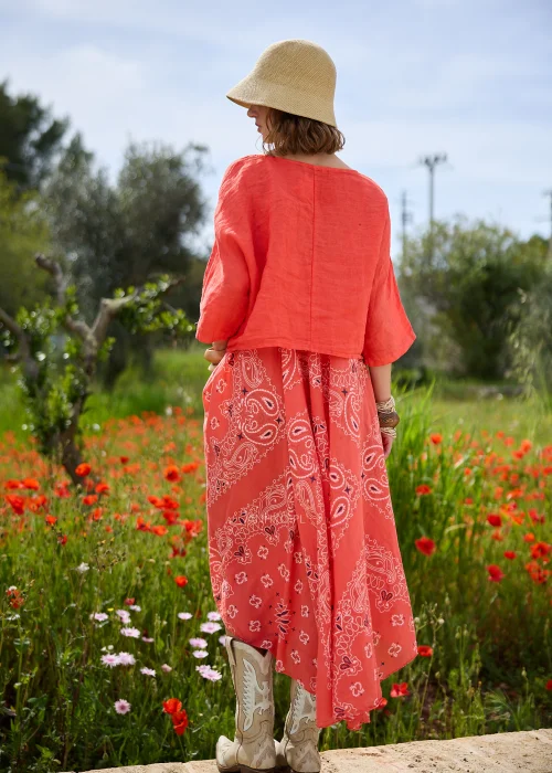 Koralowa sukienka maxi na cienkich ramiączkach z ozdobnym wzorem paisley. Zwiewny model o rozkloszowanym kroju prezentowany w stylizacji boho z kapeluszem i kowbojkami.