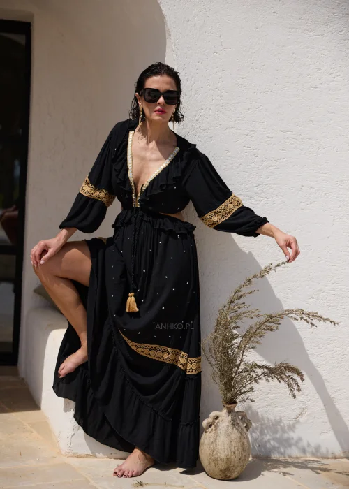 Czarna sukienka maxi w stylu boho z ażurowymi zdobieniami w kolorze złota, głębokim dekoltem V i wycięciami w talii. Zwiewny model z szerokimi rękawami i ozdobnym wiązaniem z chwostami, idealny na wakacyjne i wieczorowe stylizacje.