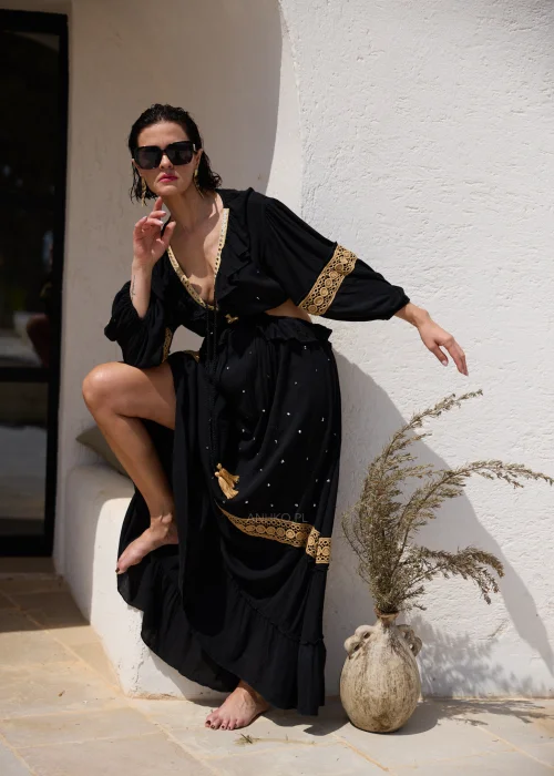 Czarna sukienka maxi w stylu boho z ażurowymi zdobieniami w kolorze złota, głębokim dekoltem V i wycięciami w talii. Zwiewny model z szerokimi rękawami i ozdobnym wiązaniem z chwostami, idealny na wakacyjne i wieczorowe stylizacje.