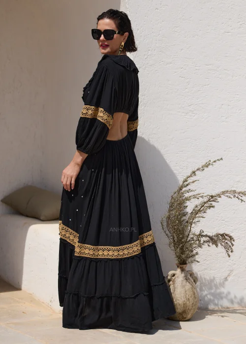 Czarna sukienka maxi w stylu boho z ażurowymi zdobieniami w kolorze złota, głębokim dekoltem V i wycięciami w talii. Zwiewny model z szerokimi rękawami i ozdobnym wiązaniem z chwostami, idealny na wakacyjne i wieczorowe stylizacje.