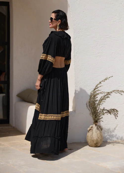 Czarna sukienka maxi w stylu boho z ażurowymi zdobieniami w kolorze złota, głębokim dekoltem V i wycięciami w talii. Zwiewny model z szerokimi rękawami i ozdobnym wiązaniem z chwostami, idealny na wakacyjne i wieczorowe stylizacje.