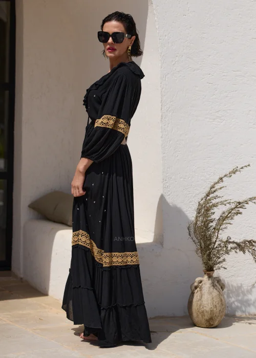 Czarna sukienka maxi w stylu boho z ażurowymi zdobieniami w kolorze złota, głębokim dekoltem V i wycięciami w talii. Zwiewny model z szerokimi rękawami i ozdobnym wiązaniem z chwostami, idealny na wakacyjne i wieczorowe stylizacje.