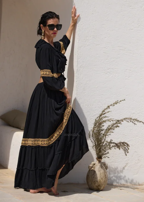 Czarna sukienka maxi w stylu boho z ażurowymi zdobieniami w kolorze złota, głębokim dekoltem V i wycięciami w talii. Zwiewny model z szerokimi rękawami i ozdobnym wiązaniem z chwostami, idealny na wakacyjne i wieczorowe stylizacje.