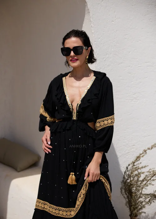 Czarna sukienka maxi w stylu boho z ażurowymi zdobieniami w kolorze złota, głębokim dekoltem V i wycięciami w talii. Zwiewny model z szerokimi rękawami i ozdobnym wiązaniem z chwostami, idealny na wakacyjne i wieczorowe stylizacje.
