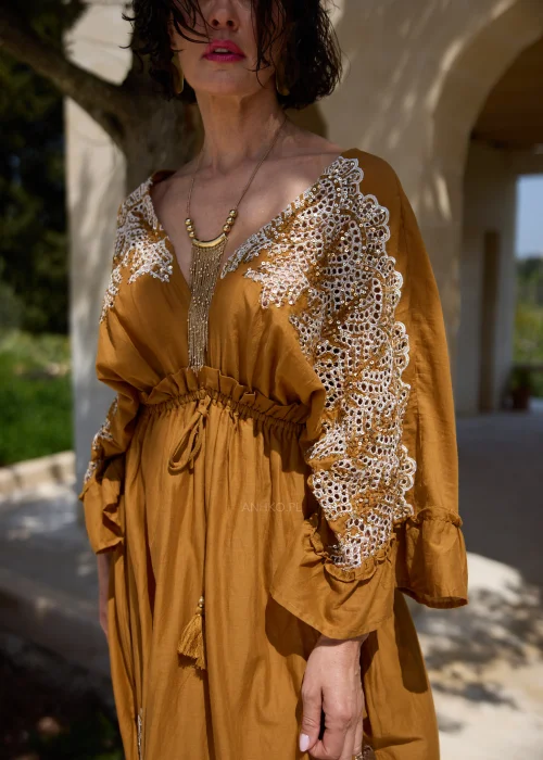 Karmelowa sukienka boho midi z ozdobną koronką i haftowanymi wstawkami przy dekolcie, rękawach oraz dole. Kobiecy fason z wiązaniem w talii i falbaną tworzy elegancką stylizację w klimacie śródziemnomorskim.