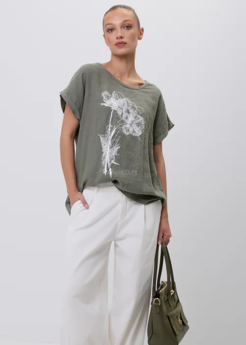 Oliwkowy damski T-shirt oversize z białym kwiatowym nadrukiem i podwijanymi rękawami. Luźna bluzka w naturalnym stylu, zestawiona z białymi szerokimi spodniami i torebką, idealna do codziennych stylizacji.