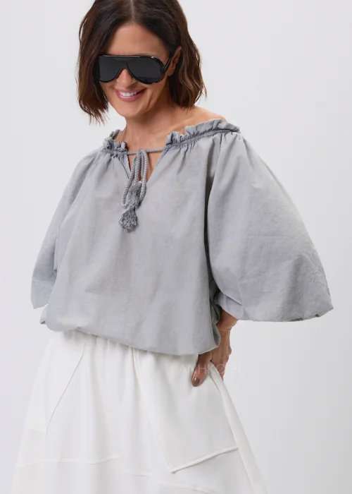 Szara bluzka damska o luźnym fasonie z bufiastymi rękawami i wiązaniem przy dekolcie zakończonym chwostami. Lekki model w stylu boho, idealny do spódnic i spodni w codziennych oraz eleganckich stylizacjach.