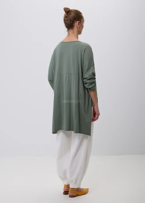 Oliwkowa damska bluzka oversize z długim rękawem i asymetrycznym dołem, wykonana z miękkiej dzianiny. Minimalistyczna stylizacja casual z białymi spodniami, idealna na co dzień i do nowoczesnych zestawów.