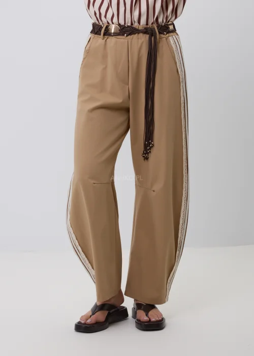 Beżowe damskie spodnie o luźnym fasonie z szerokimi nogawkami i ozdobnym koronkowym lampasem po bokach. Stylowy model casual z elastycznym pasem, szlufkami i plecionym paskiem w odcieniach brązu.