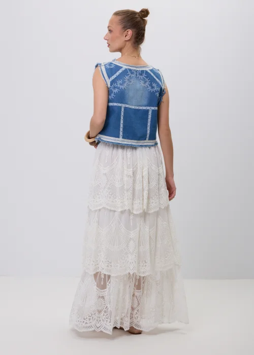 Niebieska jeansowa kamizelka damska z ozdobnym białym haftem i postrzępionym wykończeniem. Model zapinany na guziki, inspirowany stylem boho i vintage, zestawiony z koronkową spódnicą maxi w eleganckiej stylizacji.
