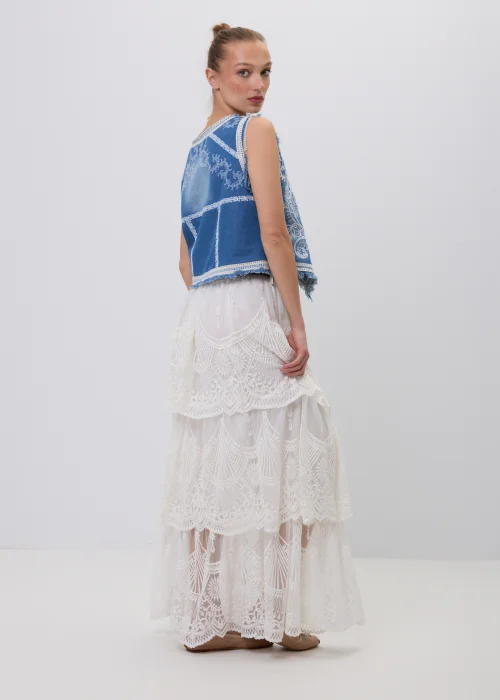 Niebieska jeansowa kamizelka damska z ozdobnym białym haftem i postrzępionym wykończeniem. Model zapinany na guziki, inspirowany stylem boho i vintage, zestawiony z koronkową spódnicą maxi w eleganckiej stylizacji.
