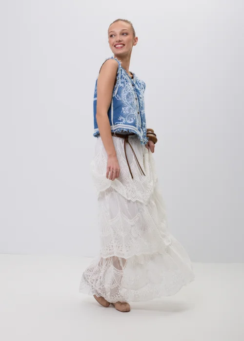 Niebieska jeansowa kamizelka damska z ozdobnym białym haftem i postrzępionym wykończeniem. Model zapinany na guziki, inspirowany stylem boho i vintage, zestawiony z koronkową spódnicą maxi w eleganckiej stylizacji.
