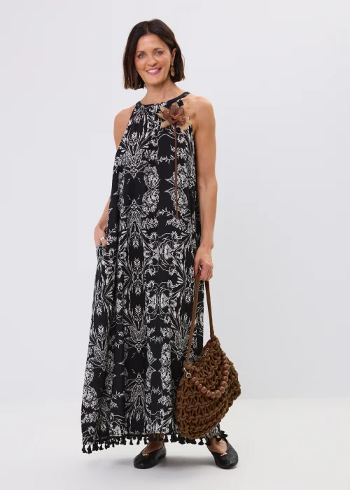Czarna sukienka maxi damska z białym roślinnym wzorem, dekoltem halter i ozdobnymi chwostami przy dole. Zwiewny fason w stylu boho uzupełniono dekoracyjnym kwiatem przy ramieniu oraz plecioną torebką.