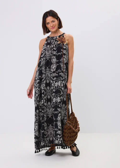 Czarna sukienka maxi damska z białym roślinnym wzorem, dekoltem halter i ozdobnymi chwostami przy dole. Zwiewny fason w stylu boho uzupełniono dekoracyjnym kwiatem przy ramieniu oraz plecioną torebką.