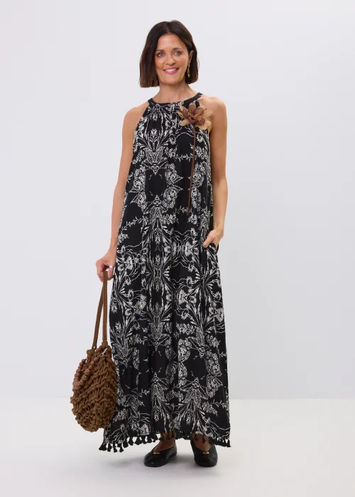 Czarna sukienka maxi damska z białym roślinnym wzorem, dekoltem halter i ozdobnymi chwostami przy dole. Zwiewny fason w stylu boho uzupełniono dekoracyjnym kwiatem przy ramieniu oraz plecioną torebką.