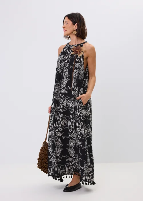 Czarna sukienka maxi damska z białym roślinnym wzorem, dekoltem halter i ozdobnymi chwostami przy dole. Zwiewny fason w stylu boho uzupełniono dekoracyjnym kwiatem przy ramieniu oraz plecioną torebką.