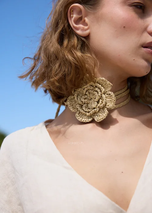 Pleciony dodatek z rafii w naturalnym beżowym odcieniu z dużym kwiatem, który można nosić jako naszyjnik lub pasek. Wielofunkcyjny model w stylu boho efektownie uzupełnia sukienki, tuniki i letnie stylizacje.