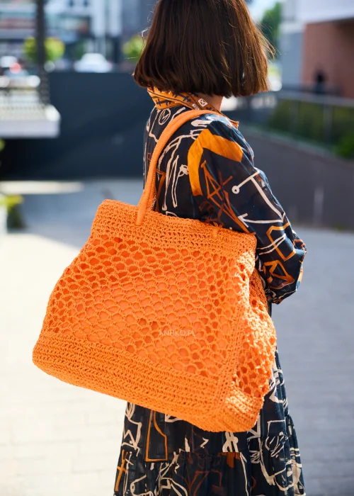Pomarańczowa ażurowa torebka shopper wykonana z plecionego splotu z krótkimi uchwytami. Lekki, pojemny model doskonale uzupełnia letnią stylizację z czarną sukienką we wzory w odcieniach pomarańczu i beżu.