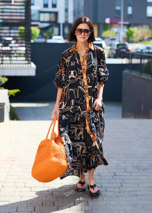Pomarańczowa ażurowa torebka shopper wykonana z plecionego splotu z krótkimi uchwytami. Lekki, pojemny model doskonale uzupełnia letnią stylizację z czarną sukienką we wzory w odcieniach pomarańczu i beżu.