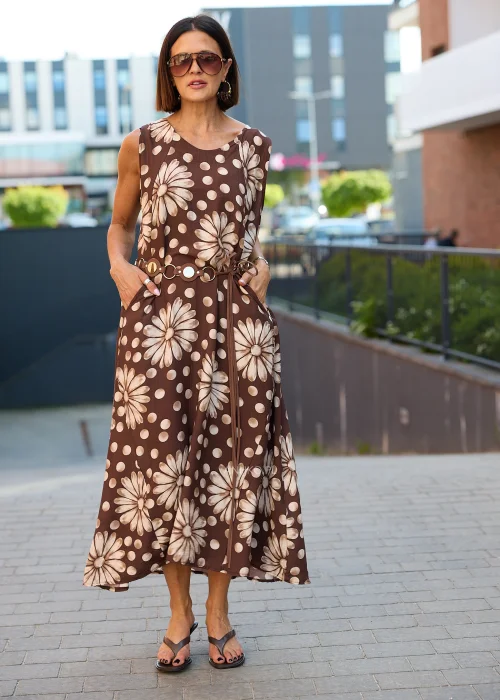 Sukienka maxi Plus Size w brązowym kolorze z dużym kwiatowym wzorem i białymi groszkami, z okrągłym dekoltem, kieszeniami i ozdobnym paskiem w talii, prezentowana w miejskiej stylizacji.