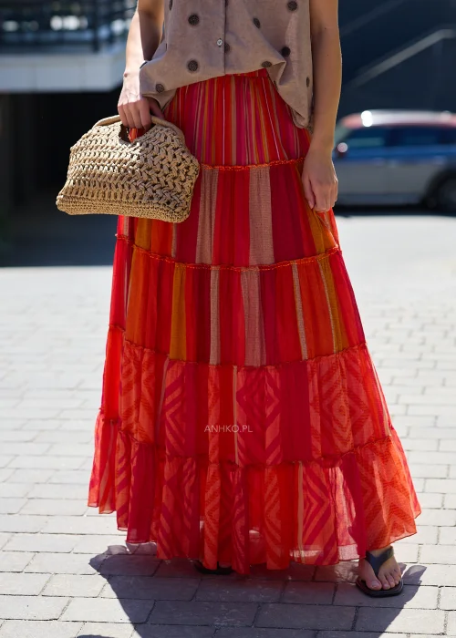 Spódnica maxi z falbanami w odcieniach czerwieni, różu i pomarańczu z patchworkowym wzorem, zestawiona z plecioną torebką w stylu boho.