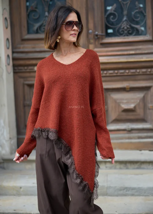 Rdzawy sweter damski oversize z dekoltem V, asymetrycznym dołem wykończonym koronką i długimi rękawami, zestawiony z brązowymi spodniami.