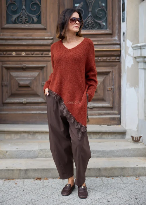 Rdzawy sweter damski oversize z dekoltem V, asymetrycznym dołem wykończonym koronką i długimi rękawami, zestawiony z brązowymi spodniami.