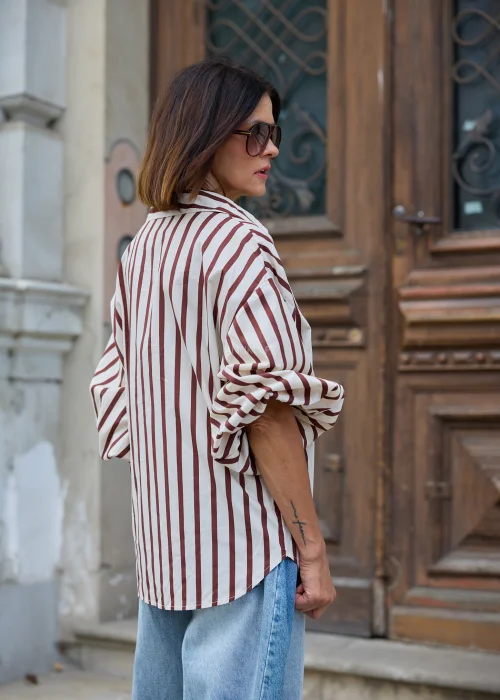 Beżowo-brązowa koszula damska oversize w pionowe paski, z klasycznym kołnierzykiem i długimi rękawami, zestawiona z jasnymi jeansami wide leg.